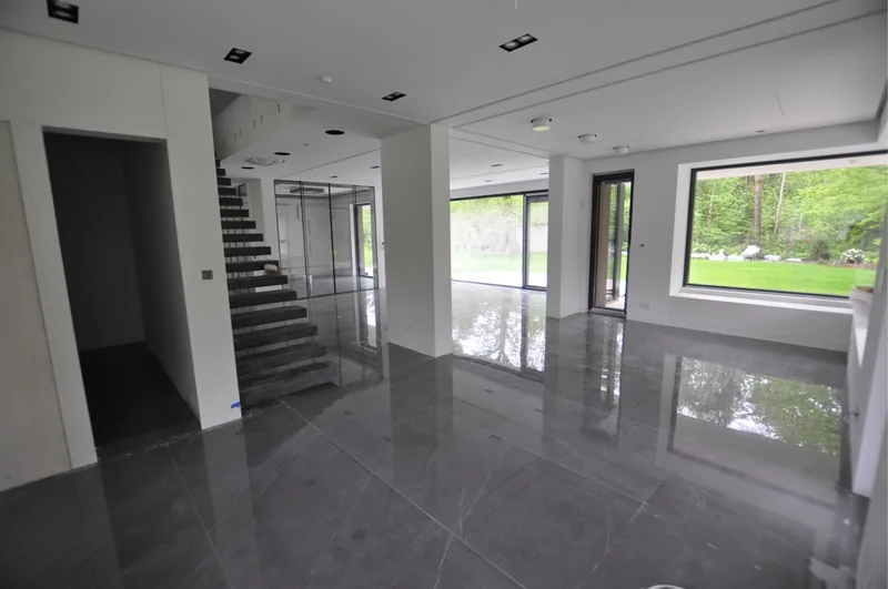 Wnętrze otwartego salonu z ciemną granitową podłogą, schodami z balustradą szklaną i widokiem na zielony ogród przez panoramiczne okna