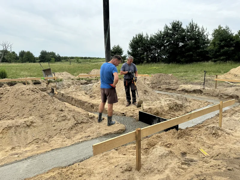 Dwóch robotników w trakcie zalewania ław fundamentowych betonem, drewniane szalunki na piaszczystej działce