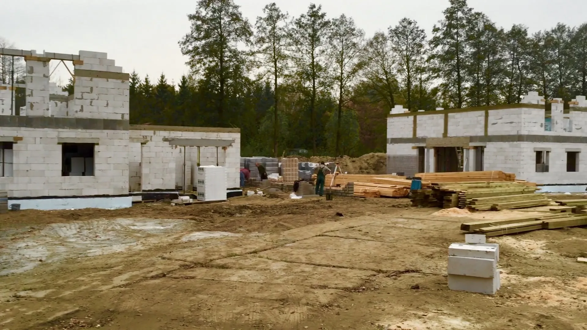 Plac budowy dwóch domów jednorodzinnych w stanie surowym otwartym — ściany z bloczków gazobetonowych, składowane drewno na więźbę, jesienna pora